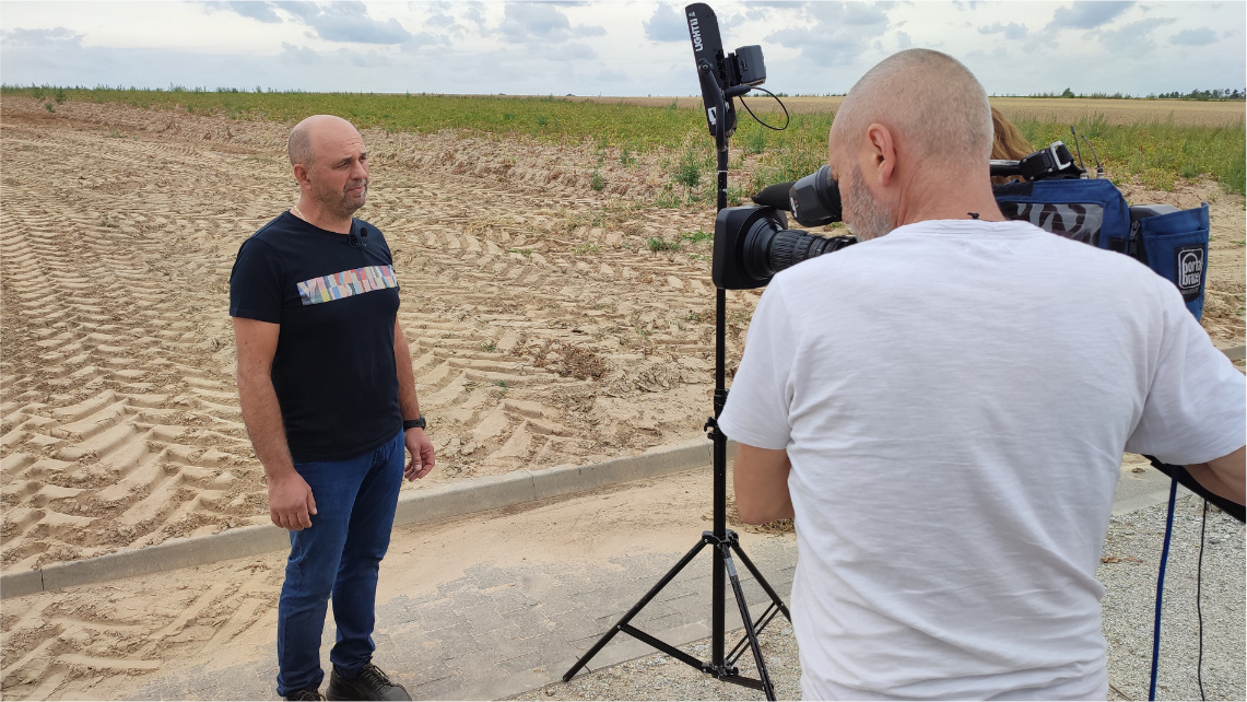 Pochmurny dzień. Remigiusz Szczepański stoi na drodze przy polu. Przed nim jest operator z kamerą i oświetlenie.