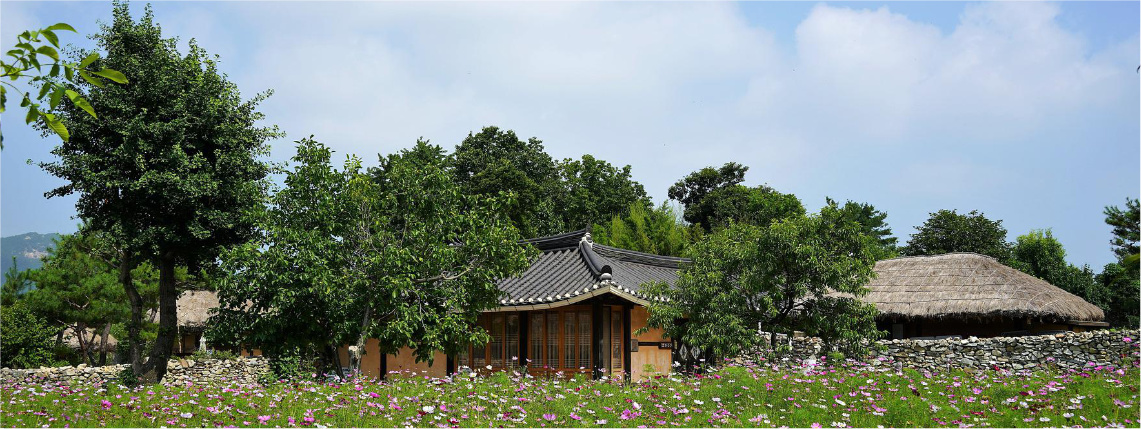 Słoneczny dzień. Na zdjęciu widoczny jest drewniany dom stojący na polu, między drzewami.