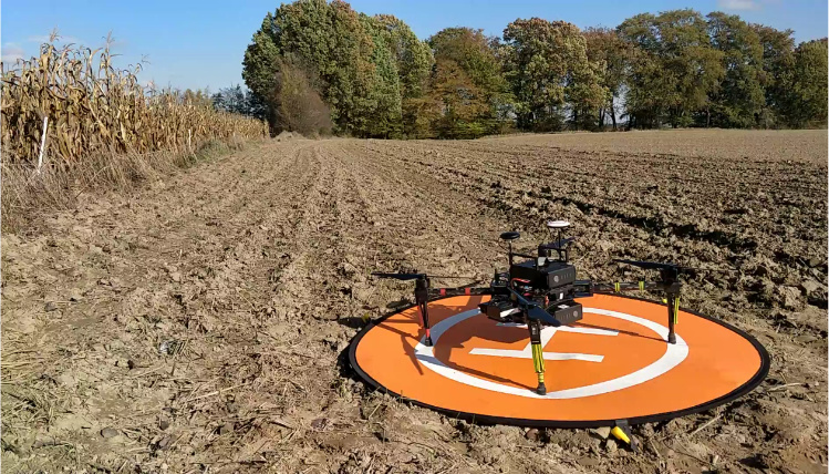 Słoneczny dzień. Na polu, w pobliżu kukurydzy, na niewielkim lądowisku stoi dron. W tle widać drzewa.