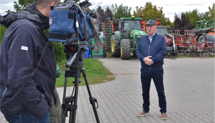 Mężczyzna na dalszym planie przemawia do kamery, która jest na bliższym planie. Obok kamery stoi operator, widoczny jest od boku. Za przemawiającym mężczyzną w tle znajdują się maszyny rolnicze.