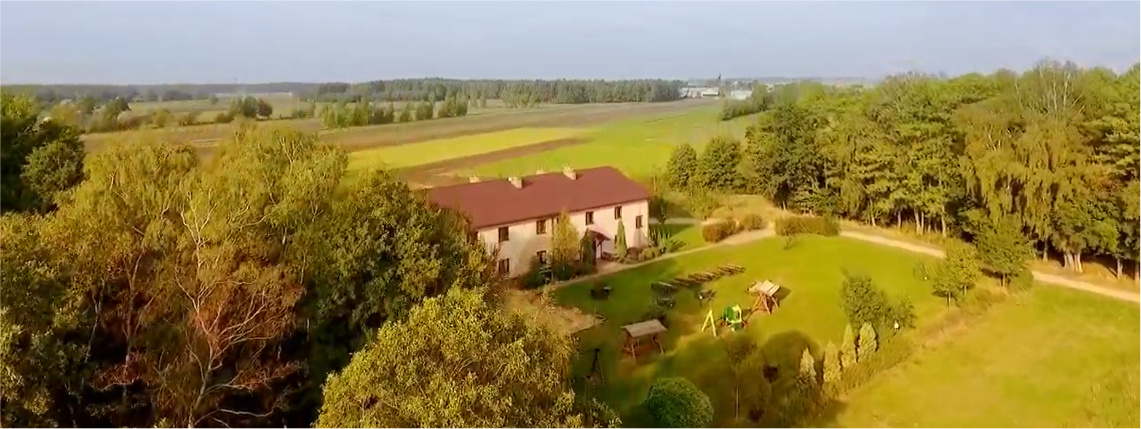 Słoneczny dzień. Z lotu ptaka widoczne jest gospodarstwo agroturystyczne, które stoi na polu, między drzewami.