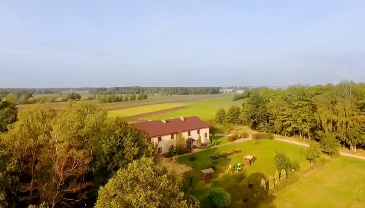 Słoneczny dzień. Z lotu ptaka widoczne jest gospodarstwo agroturystyczne, które stoi na polu, między drzewami.