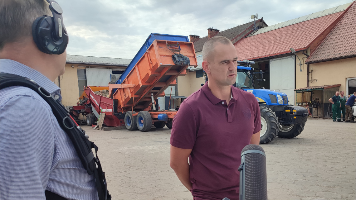 Na podwórku w gospodarstwie stoi mężczyzna, który wypowiada się do mikrofonu. Mikrofon trzyma dźwiękowiec z telewizji. W tle widać budynek oraz maszynę rolniczą.