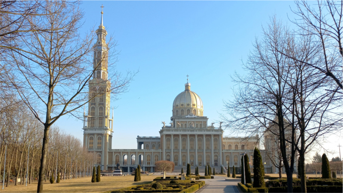 Ujęcie obejmujące całą bazylikę w Licheniu. Wieńczy ją duża kopuła i strzelista wieża, front wypełniają wysokie kolumny. Do bazyliki prowadzi aleja otoczona krzewami.