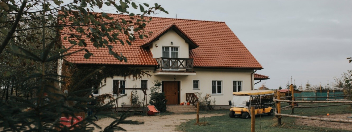 Biały dom z czerwonym dachem. Przed wejściem stoi huśtana ławka, obok na trawniku jest zaparkowany melex.