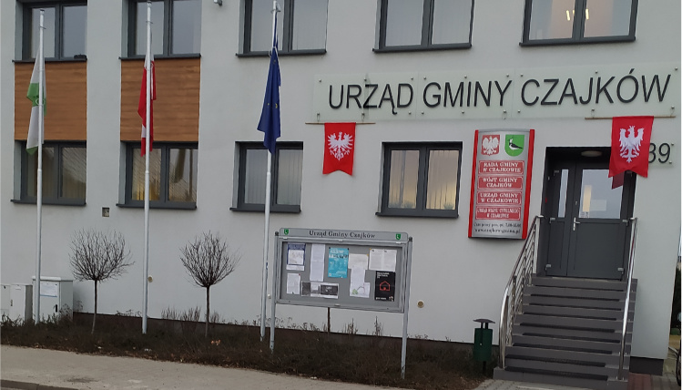 Wejście do budynku Urzędu Gminy Czajków. Na białej elewacji wiszą czerwone, urzędowe tablice, a do drzwi prowadzą schody.