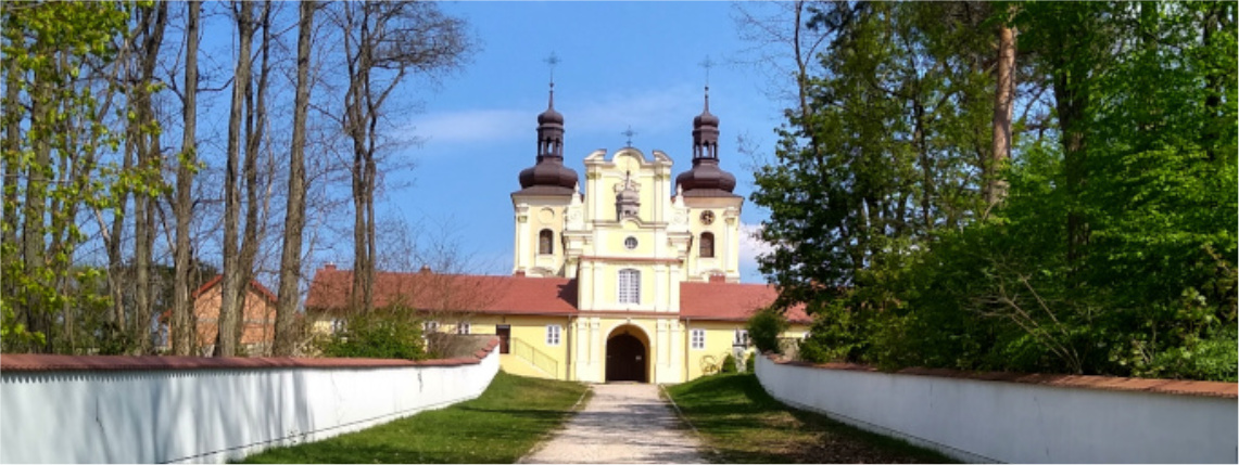 Z oddali widać kościół z dwiema wieżyczkami. Prowadzi do niego ścieżka, po obu jej stronach jest murek, a za nim drzewa.