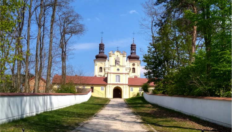 W Kazimierzu Biskupim można odwiedzić wiele miejsc Z oddali widać kościół z dwiema wieżyczkami. Prowadzi do niego ścieżka, po obu jej stronach jest murek, a za nim drzewa.