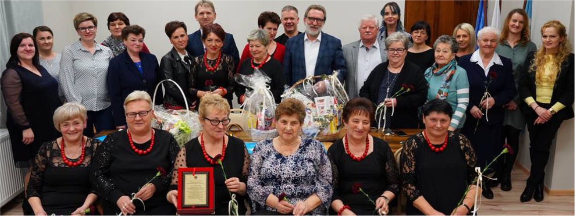 Dwa stowarzyszenia w gminie Brudzew obchodziły jubileusze istnienia W pomieszczeniu znajduje się kilkanaście osób. W pierwszym rzędzie siedzą kobiet z Koła Gospodyń Wiejskich. Jedna z nich trzyma pamiątkową tabliczkę. Na stole znajdują się kosze z prezentami.