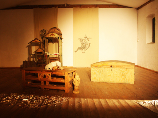 Wnętrze pomieszczenia. Znajdują się w nim drewniane meble i drewniana skrzynia. Na ścianie widać podobiznę diabła.