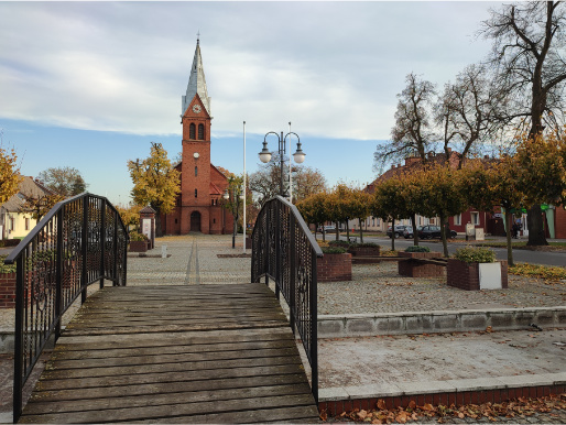 Rynek w Budzyniu. W centralnej części znajduje się drewniany mostek, a na wprost niego widać kościół z czerwonej cegły. Rynek otoczony jest drzewkami.