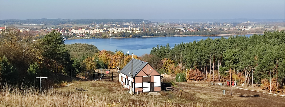 Panorama na miasto Chodzież oraz jezioro chodzieskie. Na horyzoncie widać budynki oraz lasy. W centrum zdjęcia stoi chata.