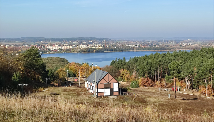 Panorama na miasto Chodzież oraz jezioro chodzieskie. Na horyzoncie widać budynki oraz lasy. W centrum zdjęcia stoi chata.