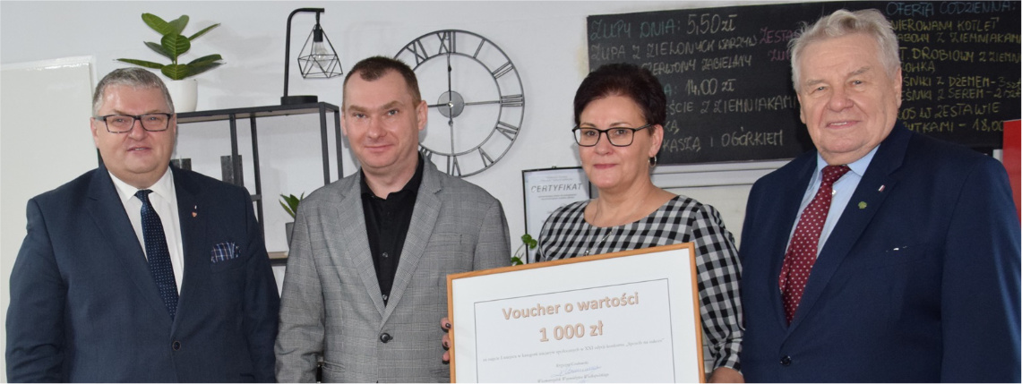 W pomieszczeniu stoją cztery osoby, trzech mężczyzn i kobieta. Kobieta trzyma dużą ramkę, w której jest voucher o wartości 1000 zł.