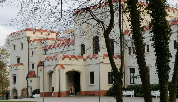 Zespół pałacowo-parkowy w Jarocinie Front pałacu o białych ścianach i kwadratowej architekturze.