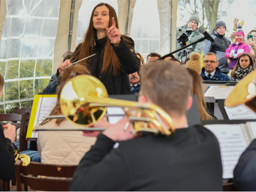 Uwaga skupiona jest na dyrygentce podczas występu orkiestry. Od tyłu widać muzyków z instrumentami. W tle znajdują się ludzie.