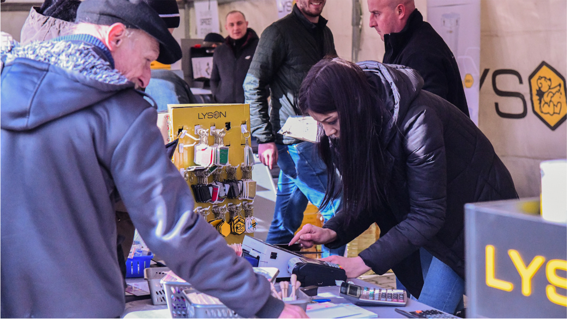 Przed straganem stoi mężczyzna, który ogląda produkty. Sprzedająca kobieta pochyla się nad kasą fiskalną.