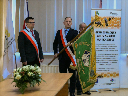 Mężczyzna z biało-czerwoną szarfą trzyma sztandar Wojewódzkiego Związku Pszczelarzy. Dwaj mężczyźni stoją po obu jego stronach.