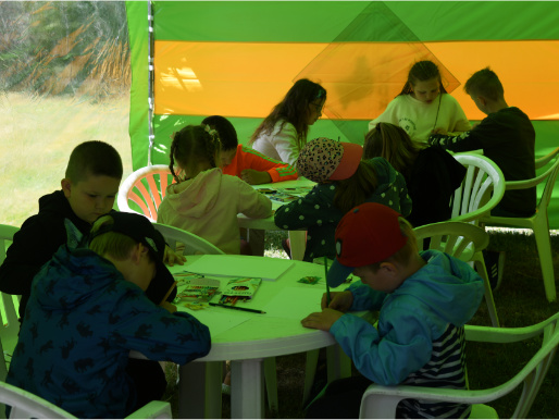 Przy stole pod namiotem siedzą dzieci i rysują.
