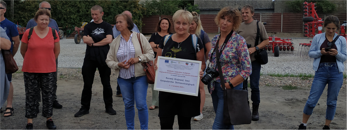 Wyjazd studyjny Krajowej Sieci Gospodarstw Demonstracyjnych do województwa kujawsko-pomorskiego Dwie kobiety pozują do zdjęcia, jedna trzyma tabliczkę informującą o operacji. Obok stoją inni ludzie.