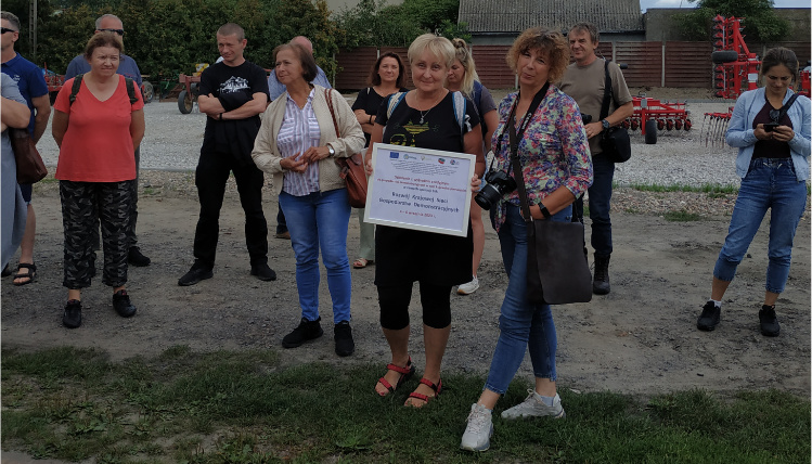 Wyjazd studyjny Krajowej Sieci Gospodarstw Demonstracyjnych do województwa kujawsko-pomorskiego Dwie kobiety pozują do zdjęcia, jedna trzyma tabliczkę informującą o operacji. Obok stoją inni ludzie.