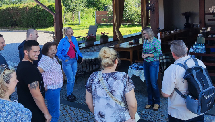 Grupa osób stoi na podwórku. Wszyscy są zwróceni w stronę kobiety, która mówi.