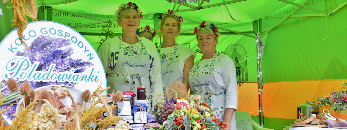 Pod namiotem stoją trzy uśmiechnięte kobiety w białych fartuchach i czerwonych wiankach na głowie. Przed nimi jest stolik z produktami oraz ozdobami z kwiatów.
