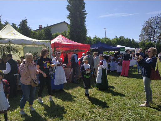 Trawiasty teren ze stoiskami pod namiotami. Przed stoiskami są ludzie.