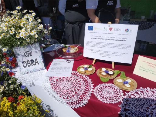 Stół pokryty obrusem, kwiatami i koronkowymi serwetkami. Na środku stoi tablica z informacją o operacji z projektu KSOW.