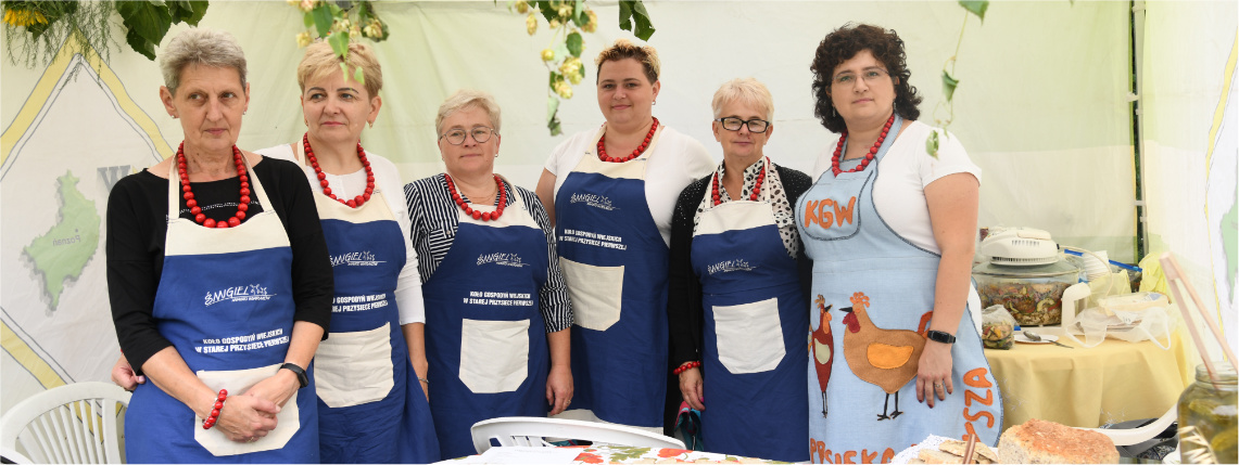  Grupa kobiet w fartuchach granatowych pozuje do zdjęcia, stojąc za stołem z potrawami.