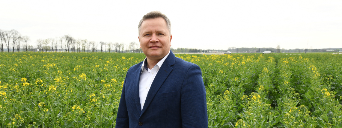 Michał Sosiński stoi wyprostowany na polu w Sielinku. W tle widoczne są uprawy.