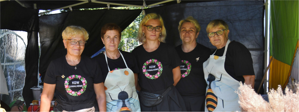 Pszczoły opanowały Gołaszyn. "Wielkopolska Wieś Zaprasza" za nami! Kobiety w fartuchach stoją przy stoisku z potrawami i pozują do zdjęcia.