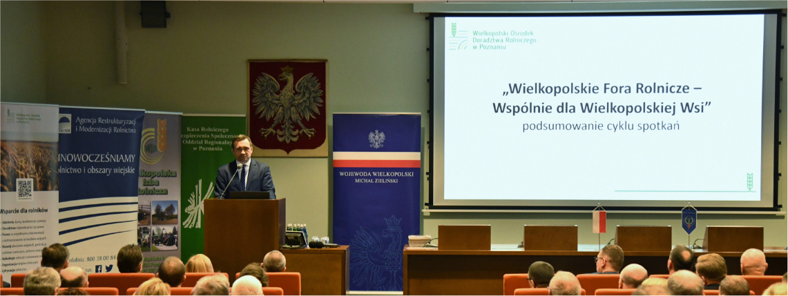 „Wielkopolskie Fora Rolnicze – Wspólnie dla Wielkopolskiej Wsi” – tysiące przeszkolonych rolników Przy mównicy stoi mężczyzna i przemawia. Obok niego jest ekran z wyświetloną prezentacją. Przed nim siedzi publiczność.